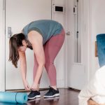 Young woman tying shoes in a bedroom, ready for a yoga session with a blue mat nearby.